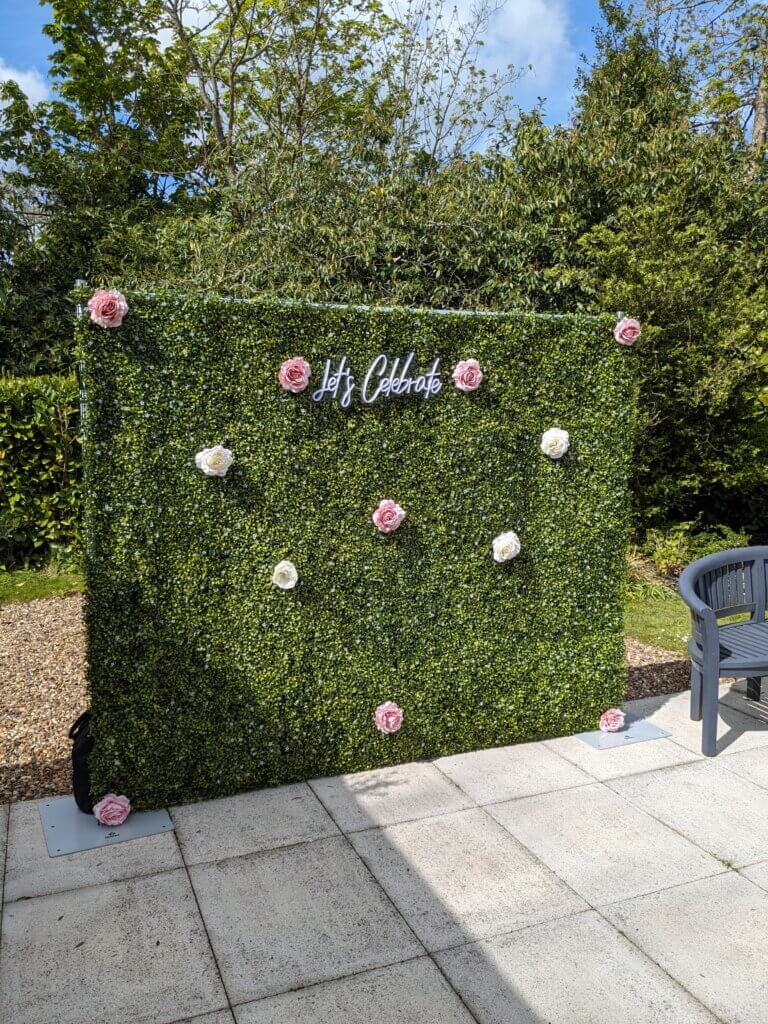 Green boxwood backdrop with pink and white rose heads, and a LED "Let's Celebrate" sign. Photographed at The Old Rectory in Padworth, Berkshire.