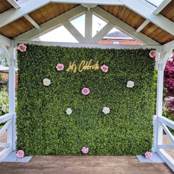 Green boxwood backdrop with LED "Let's Celebrate" sign and pink and white flower heads, displayed in wooden gazebo at Mill House Hotel