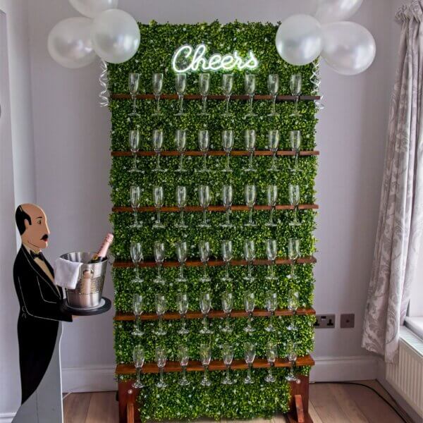 Whole green boxwood prosecco wall is on display. Filled with 48 empty prosecco glasses (champagne flutes), LED "Cheers" sign at the top and white balloons either side. Butler cutout to the side of the wall holding a champagne bucket with pink Moet champagne inside.