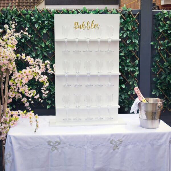 White table prosecco wall sits on top of table with white table cloth. The table prosecco wall has 3 shelves, each holding 5 glasses (15 in total). Gold vinyl at the top states "Bubbles". Champagne bucket sitting to the side with a bottle of pink Moet champagne.