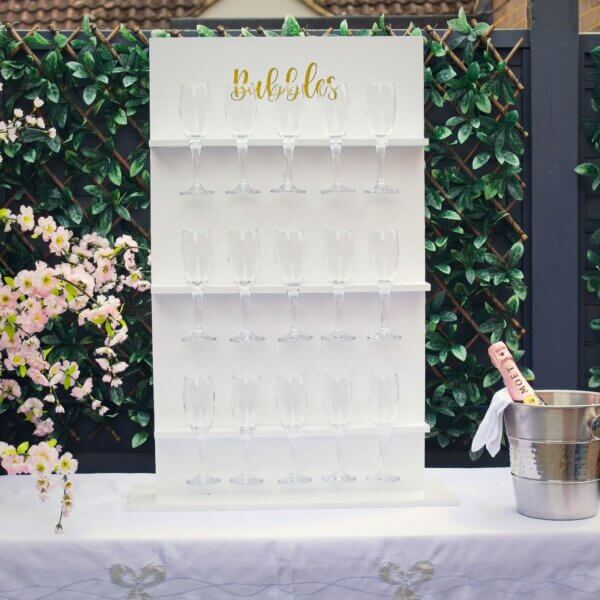 White table prosecco wall sits on top of table with white table cloth. The table prosecco wall has 3 shelves, each holding 5 glasses (15 in total). Gold vinyl at the top states "Bubbles". Champagne bucket sitting to the side with a bottle of pink Moet champagne.
