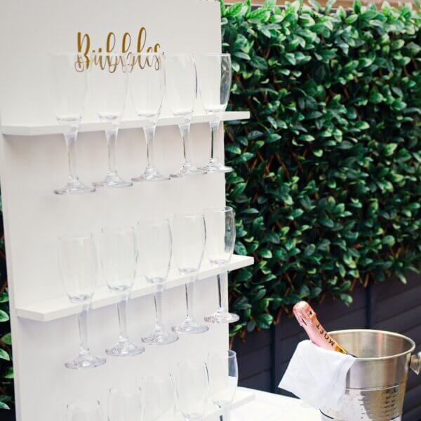 Close up and side angle of the white table prosecco wall. The top two shelves are visible and parts of the 3rd bottom shelf. All 15 glasses are just visible and wall has gold vinyl writing at the top, saying "Bubbles". A champagne bucket holding a pink Moet champagne bottle is visible to the right.