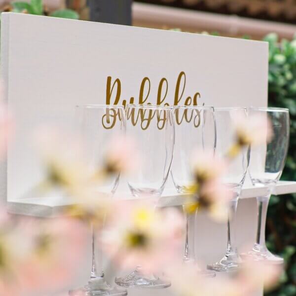 Close up of white prosecco wall, with only the top row of 5 glasses being visible. The word "Bubbles" can been seen in gold writing at the top of the wall. A blurred pink flower can be seen in front of the wall.