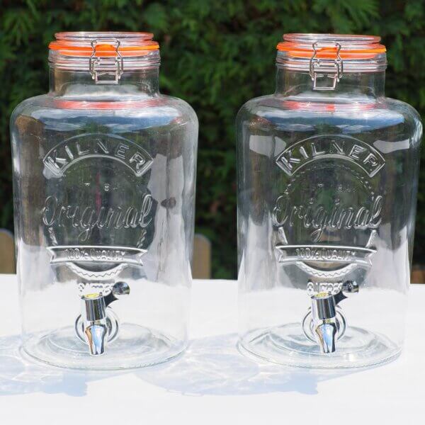 Two empty 8L Kilner drink dispensers shown sitting side by side, with faucets protruding out of the front of the large glass dispensers.