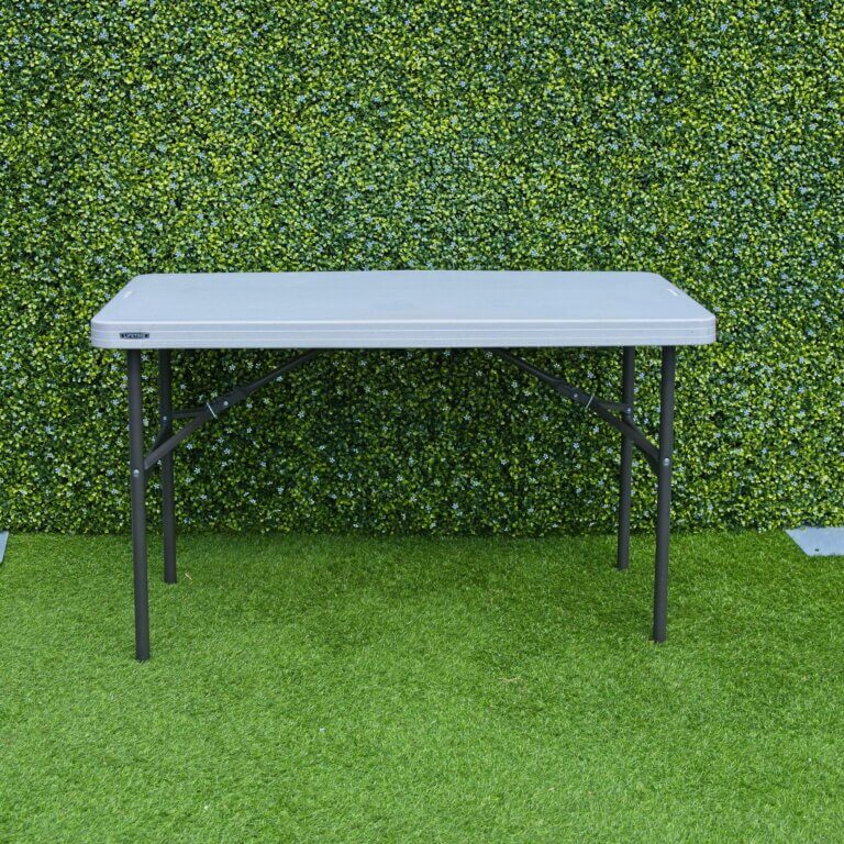 4ft plastic lifetime folding trestle table seen erected outside on grass, in front of a green boxwood backdrop