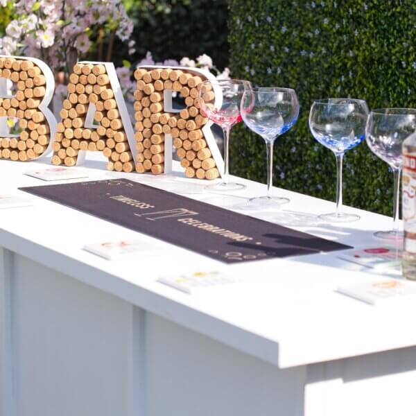 Worktop of white mobile bar visible with a portion of the front visible. On top of the bar, there is, a Bar sign made from corks, colourful gin glasses, coasters and a bar runner saying "Timeless Celebrations"