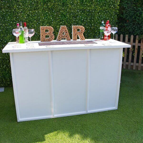 Large white mobile bar seen from the front. The worktop is visible and hosts a range of items on top of it including; a bar sign made from corks, bar runner saying "Timeless Celebrations", coasters, gin glasses and bottles of alcohol