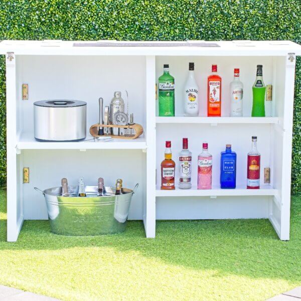 Bar of the white mobile bar is shown. The back is split into a left and right side. The left side has one shelf holding an ice bucket and cocktail utensils, with a large metal drink tub below on the floor holding bottles of champagne. The right side of the back has two shelves and both are full of alcohol bottles.