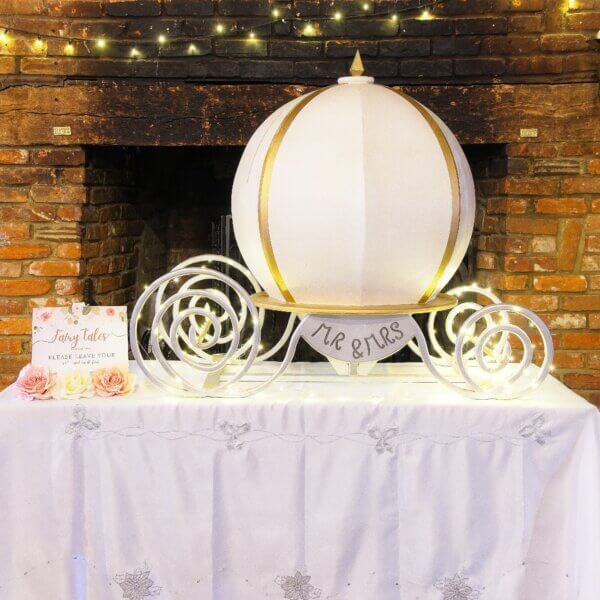 Large pumpkin style carriage post box. White with gold accents and silver carriage base and twinkle lights. Beside is a sign saying "Fairy Tales do come true. Please leave your wishes and cards here". Both of these are sitting on top of a table.