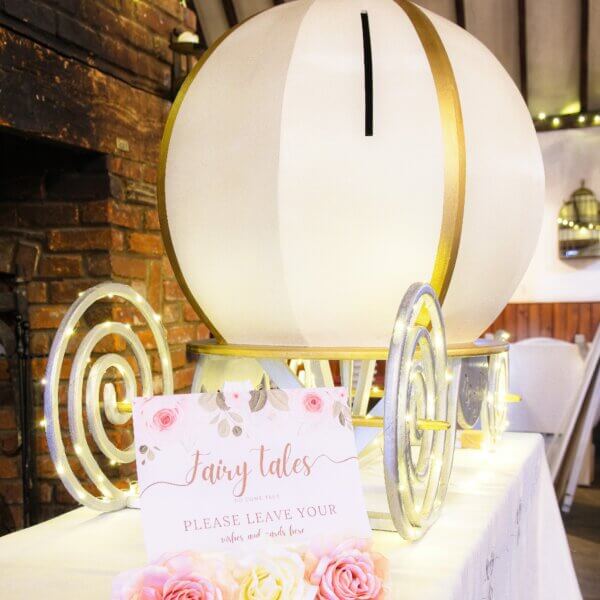 Side angle view of large pumpkin style carriage post box. White with gold accents and silver carriage base and twinkle lights. Beside is a sign saying "Fairy Tales do come true. Please leave your wishes and cards here". Both of these are sitting on top of a table.