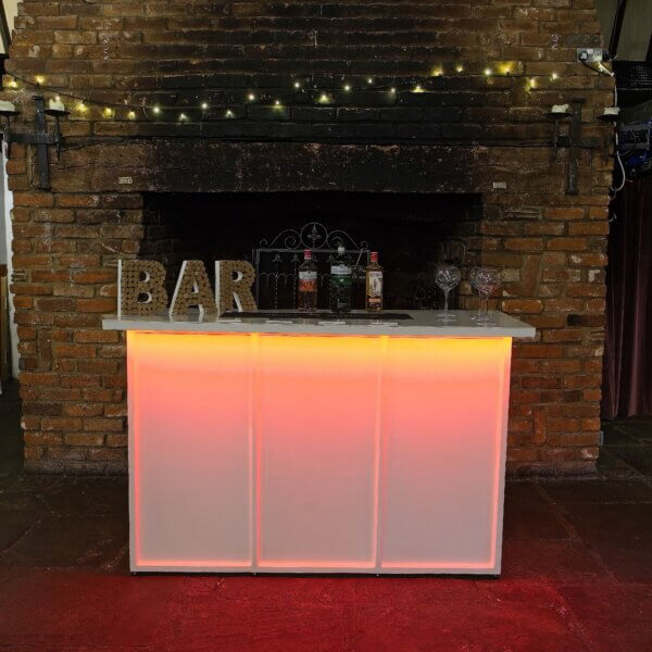 White mobile bar is shown with a bright LED light under the worktop at the front. This is shining red light down on the front of the bar. A cork bar sign, bottles of alcohol and gin glasses are shown on top of the LED bar