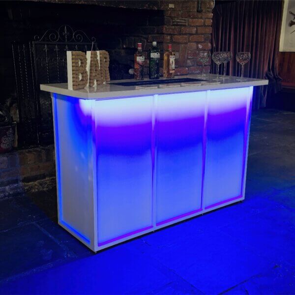 White mobile bar is shown, from an angle with a bright LED light under the worktop at the front. This is shining blue light down on the front of the bar. A cork bar sign, bottles of alcohol and gin glasses are shown on top of the LED bar