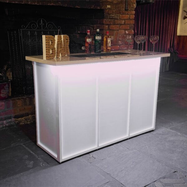 White mobile bar is shown, from an angle with a bright LED light under the worktop at the front. This is shining white light down on the front of the bar. A cork bar sign, bottles of alcohol and gin glasses are shown on top of the LED bar