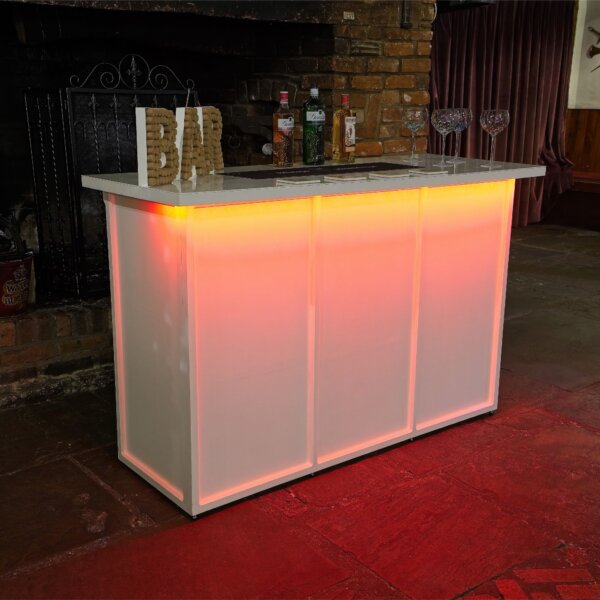 White mobile bar is shown, from an angle with a bright LED light under the worktop at the front. This is shining red light down on the front of the bar. A cork bar sign, bottles of alcohol and gin glasses are shown on top of the LED bar