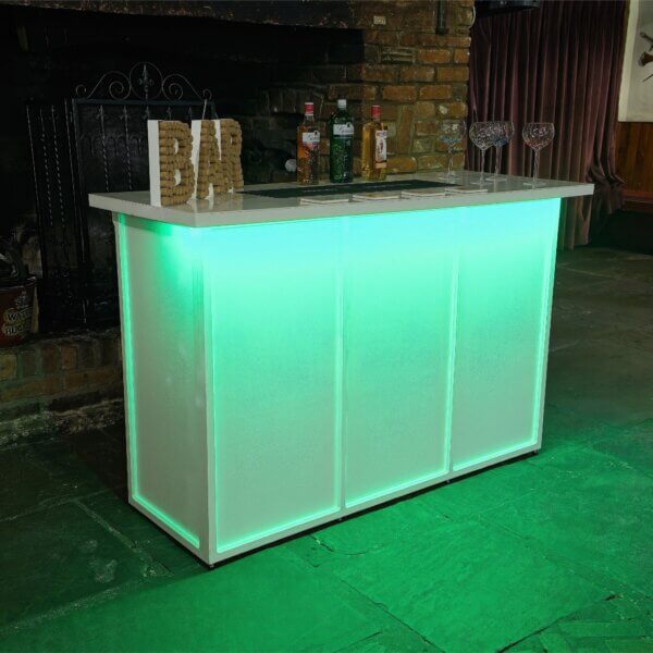 White mobile bar is shown, from an angle with a bright LED light under the worktop at the front. This is shining green light down on the front of the bar. A cork bar sign, bottles of alcohol and gin glasses are shown on top of the LED bar