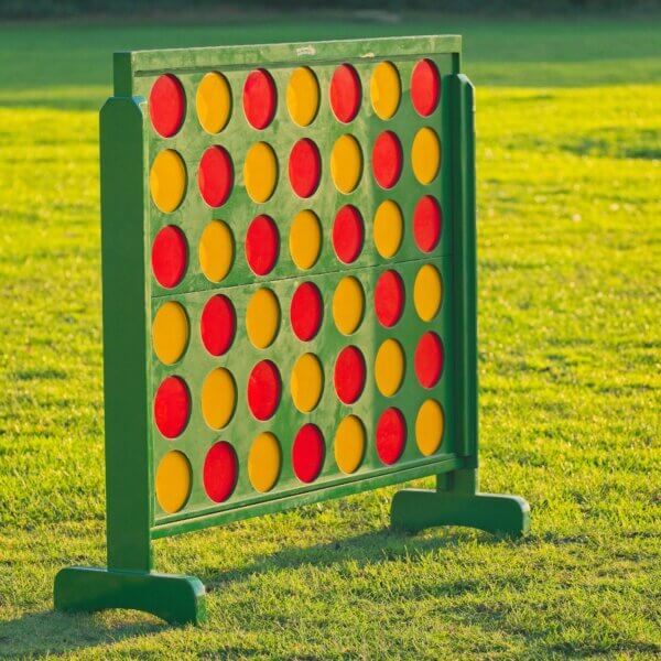 Giant connect 4 seen standing at an angle outside with the sun gleaming off of it. The frame of the connect 4 is green and filled with red and yellow counters. The counters are arranged in a red then yellow pattern.