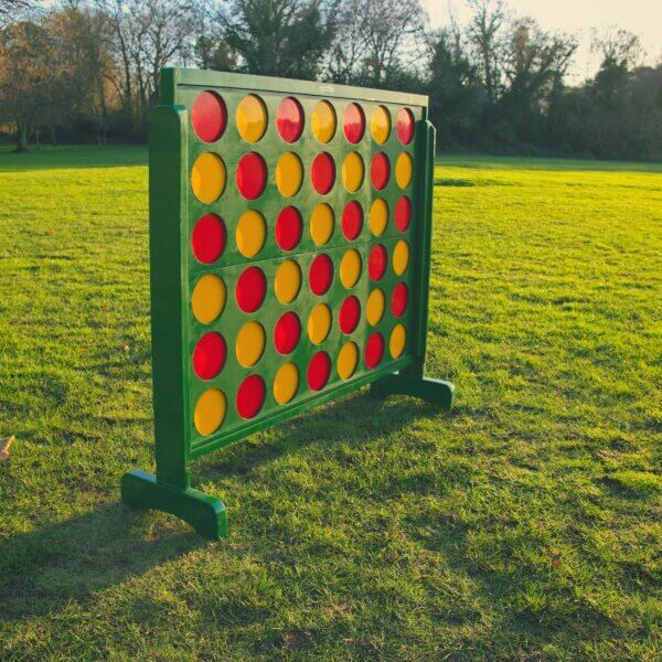 Giant connect 4 seen standing at an angle outside with the sun gleaming off of it. The frame of the connect 4 is green and filled with red and yellow counters. The counters are arranged in a red then yellow pattern.