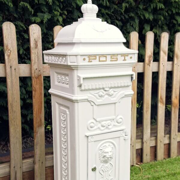 Side angle view of white ornate metal post box pictured outside. Lock shown of a front door of the post box with a embossed lions head visible, a flap for post and gold written "Post" at the top