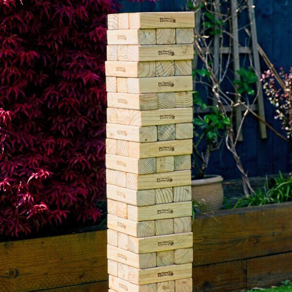 Giant Jenga tower sitting on top of a small wooden table outside. The wooden blocks are arranged 3 blocks per row and the next row is turned 90 degrees. The tower stands at 19 rows high.