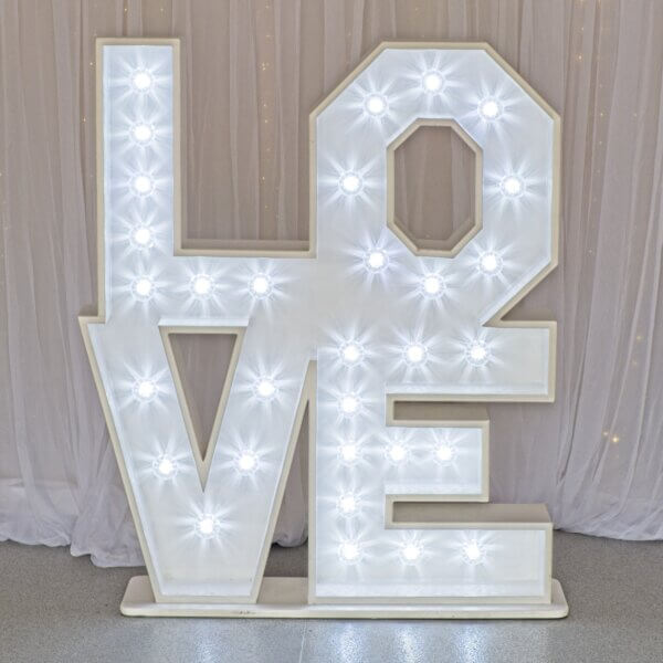 Bright white Cabochons / fairground lights are shining withing stacked LOVE marquee letters. All letters are part of one unit and the LO is sitting on top of the VE. The letters are sitting in front of white drape backdrops with curtain lights behind