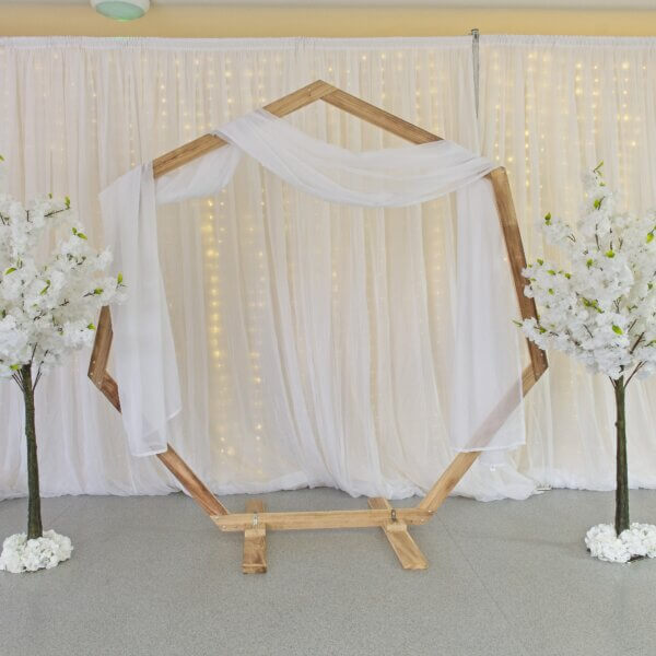 Wooden heptagonal wedding arch/arbor with white material draping over the arch. A white blossom tree stands either side of the wooden arch and all in front of a white drape backdrop with curtain lights