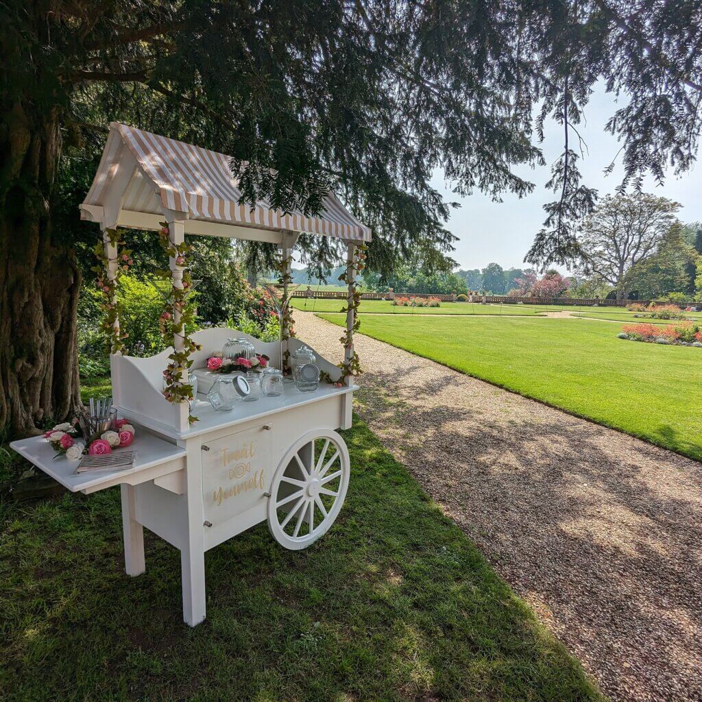 Sweet cart with gold and white top canvas, gold lettered sign on the front and gold and white sweet bags and utensils on the side table. Various sweet jars displayed. Sweet cart in in front of beautiful gardens of the Elvetham Hotel