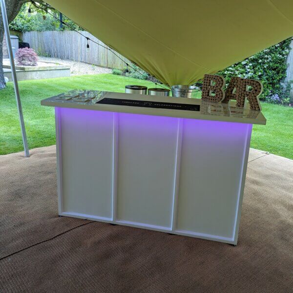 LED mobile bar shown at customer's party. A purple glow can be seen coming from the underside of the bar worktop. On top of the bar, a cork bar sign can be seen, alongside a bar runner saying "Timeless Celebrations" and coasters