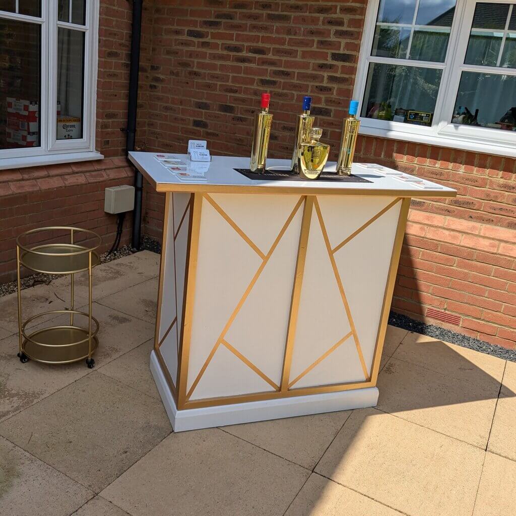 The gold and white mobile bar can be seen at a customer's party. The art deco design of the front of the bar is visible with gold lines creating geometric shapes on the white background. Gold bottles of AU vodka are sitting on the bar runner on top of the bar with a gold drinks trolley sitting beside the bar.