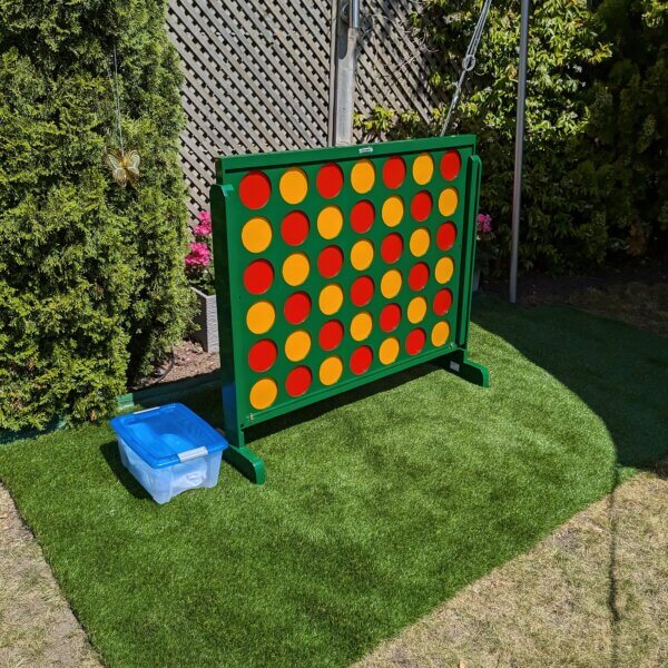 Giant connect 4 seen standing at an angle outside with the sun gleaming off of it. The frame of the connect 4 is green and filled with red and yellow counters. The counters are arranged in a red then yellow pattern.