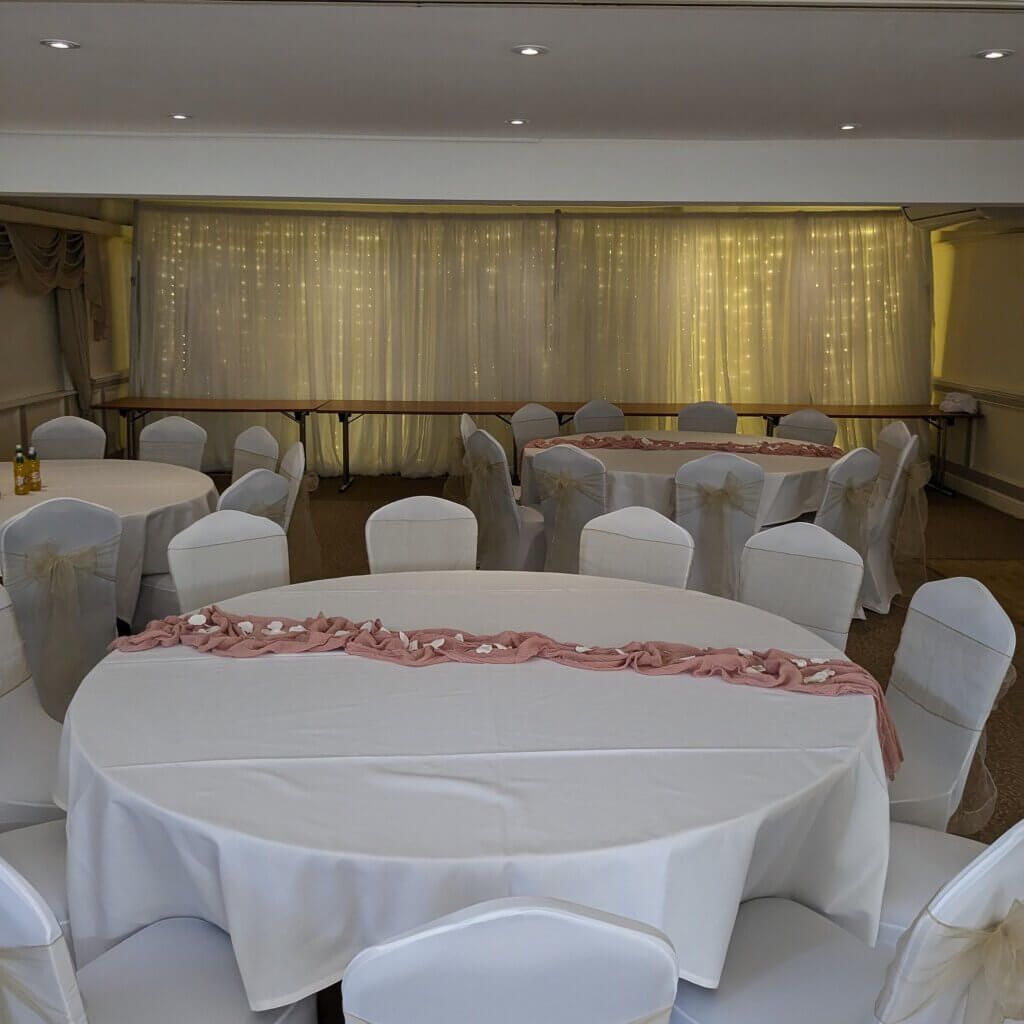 6m of white drape backdrop can be seen at the back of a reception room, at the Lismoyne Hotel, with fairy curtain lights shining through the backdrop. 3 large round tables, shown in front of the backdrop with white table cloths and white stretch chair covers.