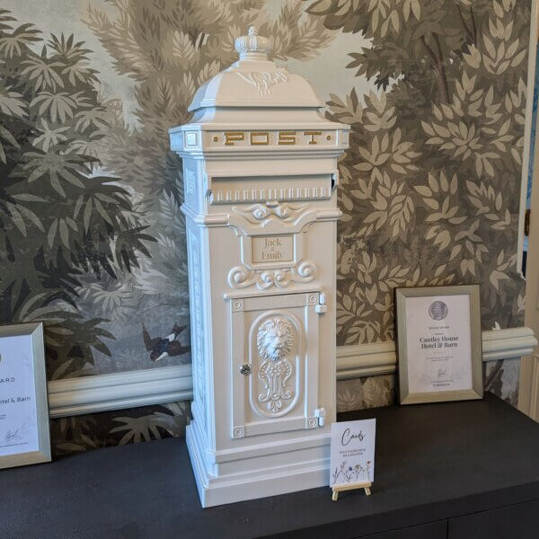 White ornate metal post box pictured on table at customer's wedding. Lock shown of a front door of the post box with a embossed lions head visible, a flap for post and gold written "Post" at the top. Gold vinyl writing displaying the couples' names is in the middle of the post box