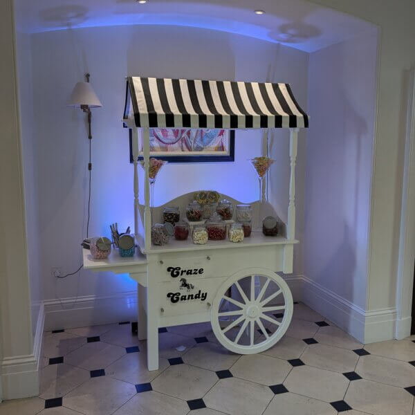 Sweet cart with black and white canvas, sign and bags. Various jars, all filled to the brim with different sweets. Custom sign on the front and LED uplights behind the sweet cart