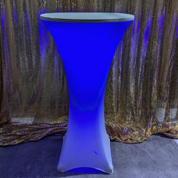 Poseur table shown with white stretch cover and blue glowing light come from within the stretch cover. Sitting in front of a gold sequin backdrop.