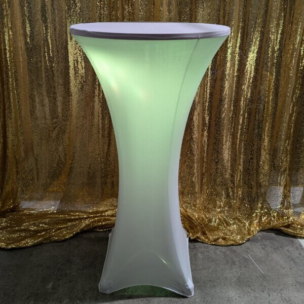 Poseur table shown with white stretch cover and yellow glowing light come from within the stretch cover. Sitting in front of a gold sequin backdrop.