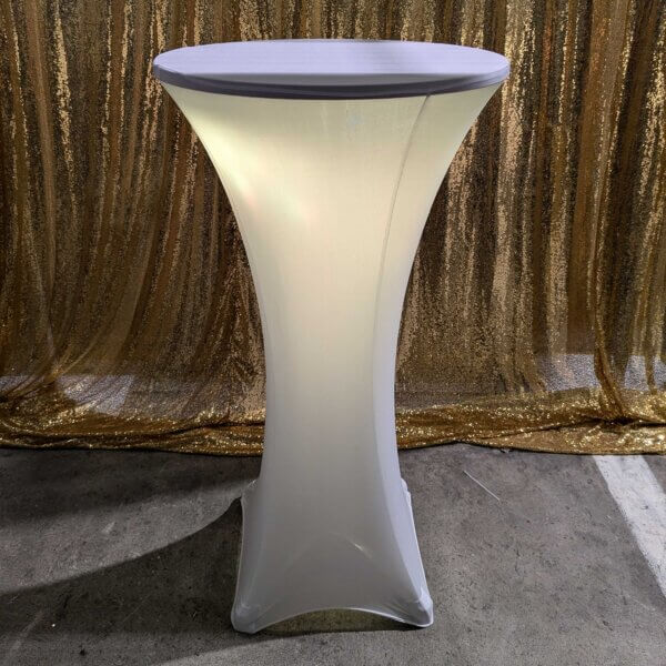 Poseur table shown with white stretch cover and white glowing light come from within the stretch cover. Sitting in front of a gold sequin backdrop.