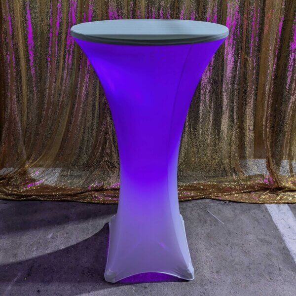 Poseur table shown with white stretch cover and purple glowing light come from within the stretch cover. Sitting in front of a gold sequin backdrop.