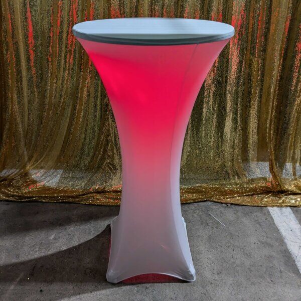 Poseur table shown with white stretch cover and red glowing light come from within the stretch cover. Sitting in front of a gold sequin backdrop.