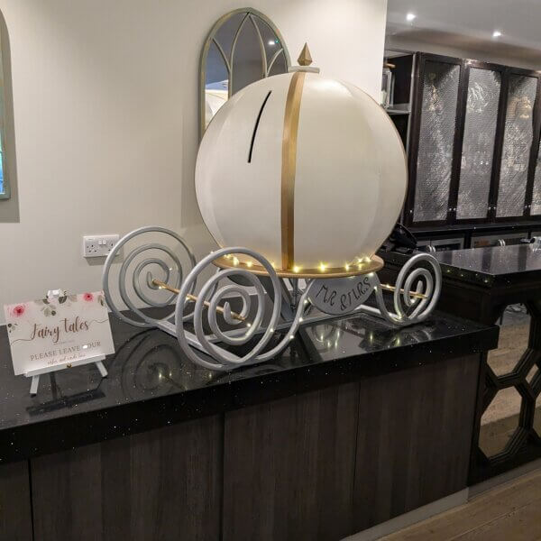 Fairy tale pumkin carriage post box sitting on worktop at Gorse Hill Hotel. The top of the carriage is white with gold accents and the base is silver covered with fairy lights. The slit for the post can be found on the top of the carriage. A sign sits next to the carriage saying "Fairy Tales do come true. Please leave your wishes and cards here".