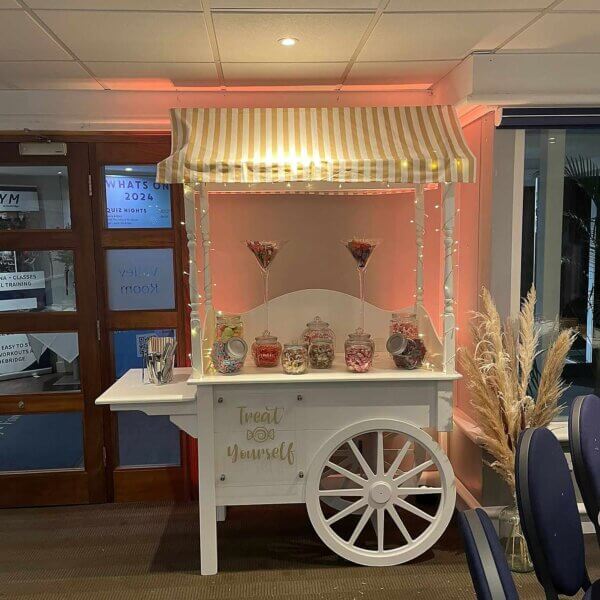 Sweet cart filled with jars and sweets, topped with gold and white striped canvas, pink vinyl lettered sign at the front. Fairy lights wrapped around the uprights of the sweet cart. LED uplights highlight the sweet cart from behind