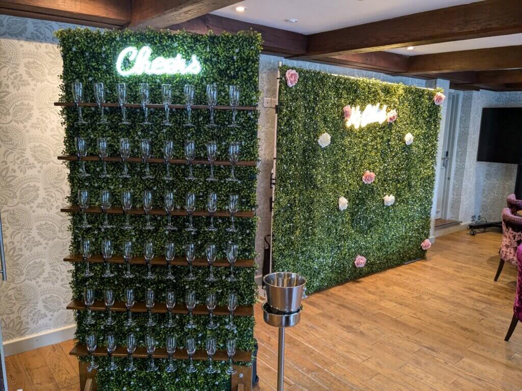 Green boxwood backdrop with pink and white flower heads sitting behind green prosecco wall with 48 prosecco glasses and LED "Cheers" sign