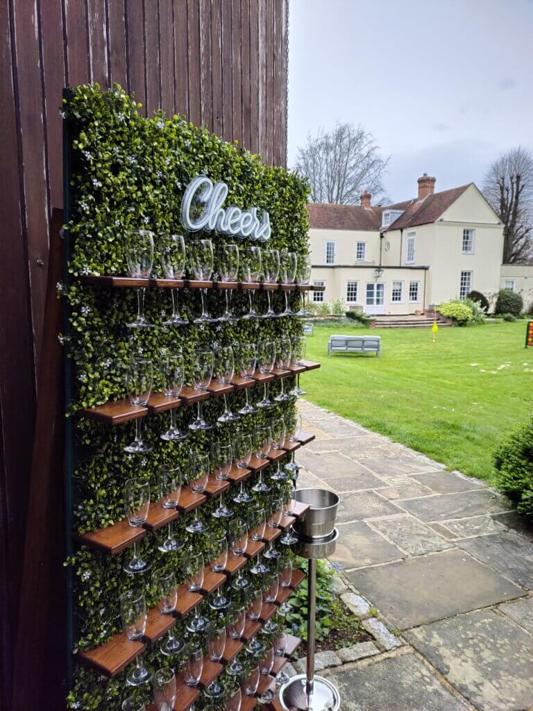 Green boxwood prosecco wall holding 48 glasses and with a LED sign at the top saying "Cheers" in front of the garden at Hillfields Farm in Pangbourne, Berkshire