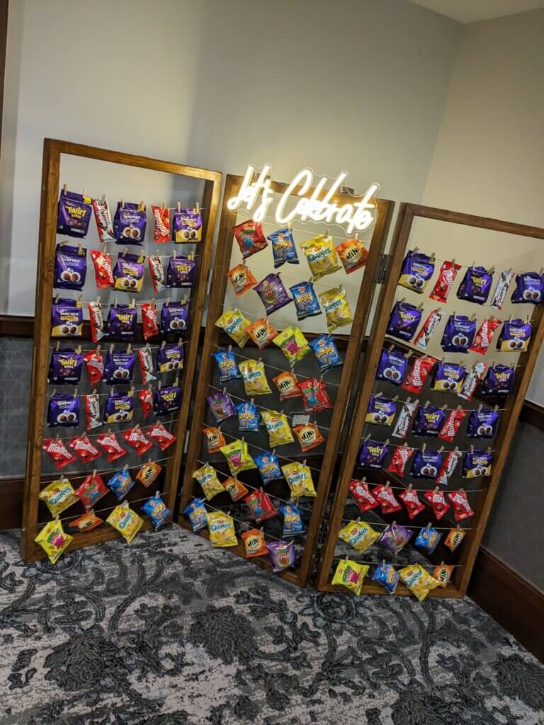 Dark wood snack wall, consisting of three concertina panels with string rows and pegs holding crisps, sweets and chocolates. A bright LED sign sits at the top of the middle panel reading "Let's Celebrate"