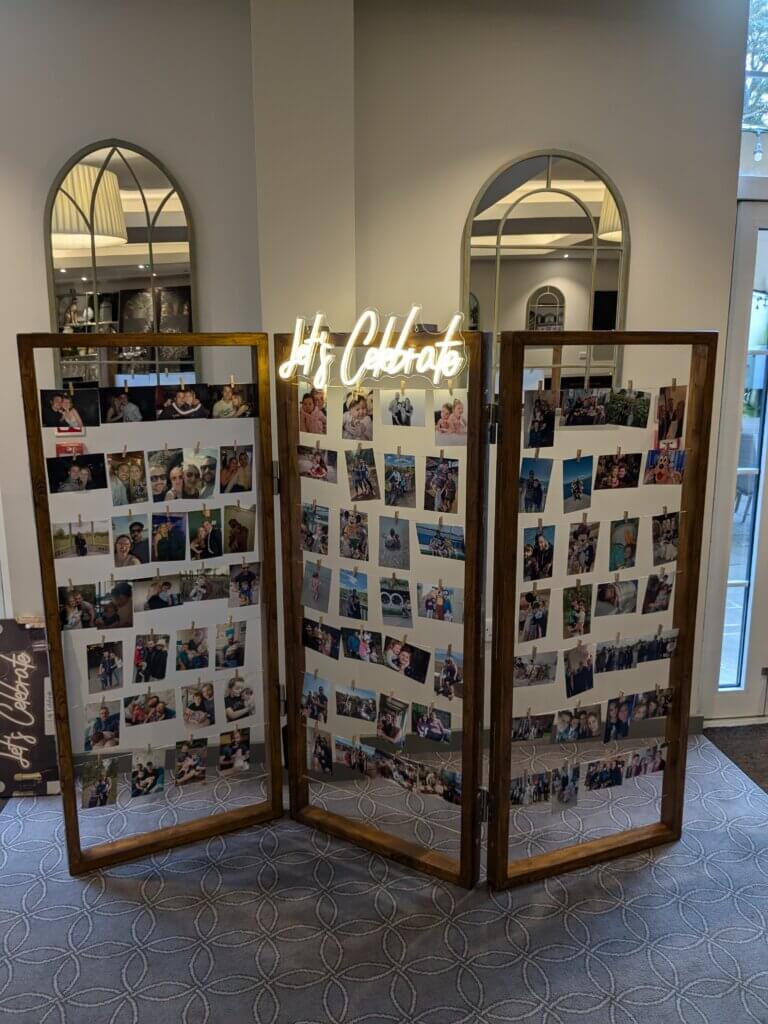 Dark wood photo wall, consisting of three concertina panels with string rows and pegs holding multiple photos. A bright LED sign sits at the top of the middle panel reading "Let's Celebrate"