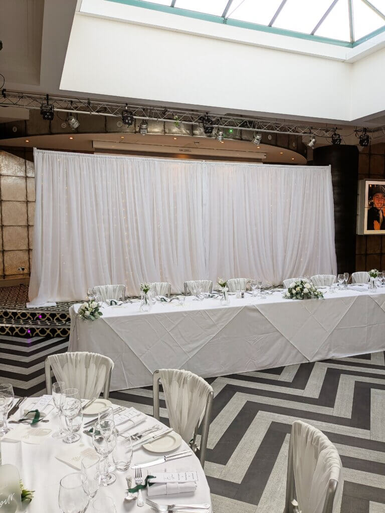 Large white drape backdrop sitting behind the head table at a wedding reception. Sparkling lights can be seen behind the drapes.