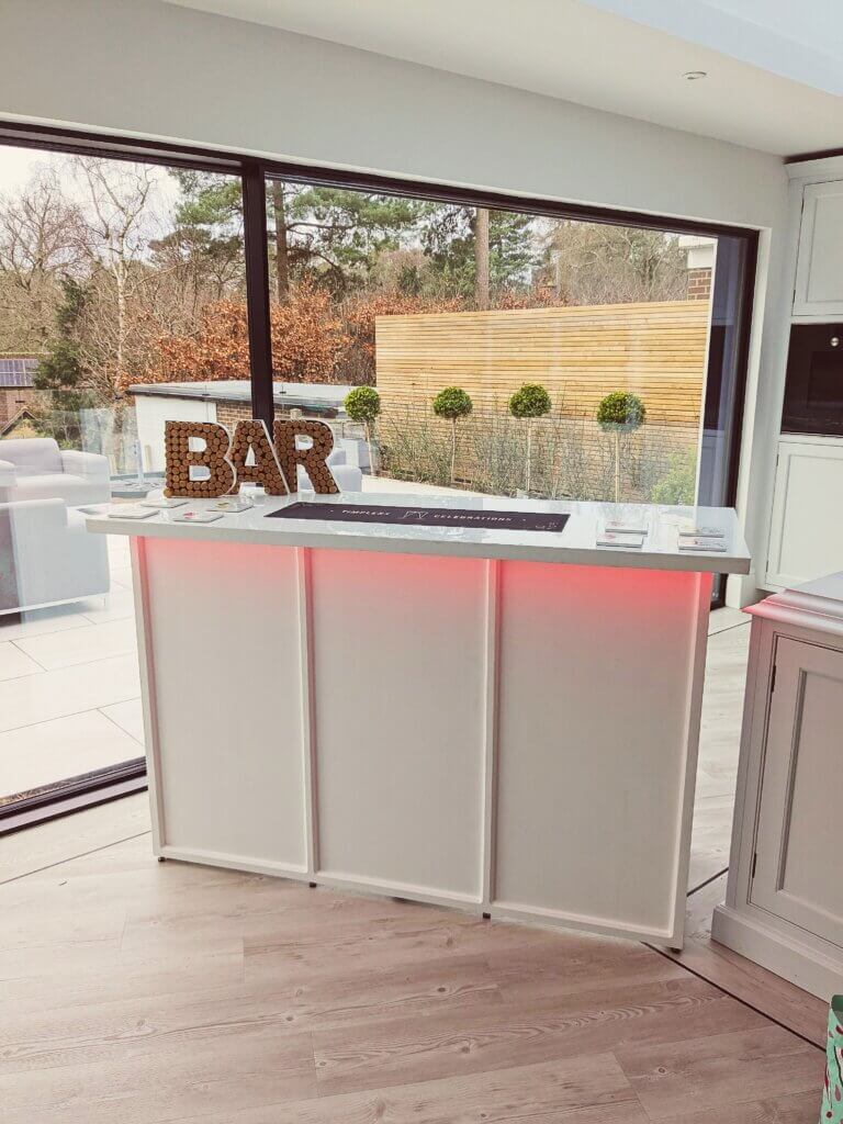 White wooden mobile bar show inside kitchen. Red light glows from the underside of the bar worktop and a bar sign sits on top of the bar top