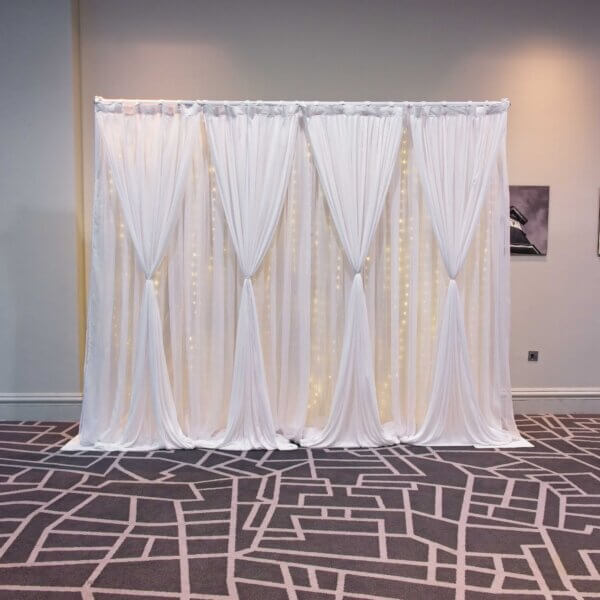 Double layered white drape backdrop with hour glass shape drapes on the front row. Curtain lights can be seen behind the drapes