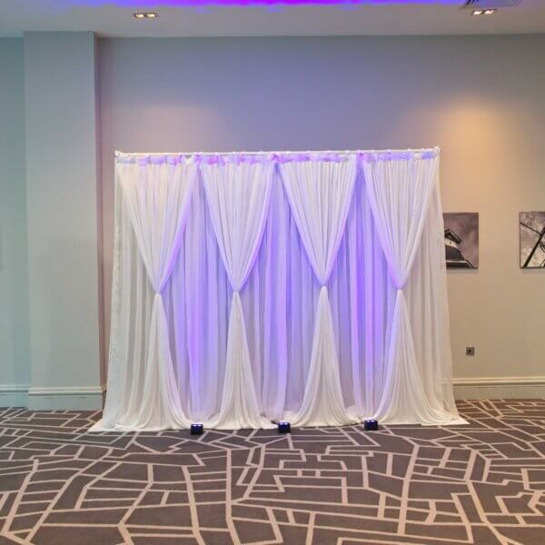 Double layered white drape backdrop with hour glass shape drapes on the front row. Purple glow from uplights can be seen on the front