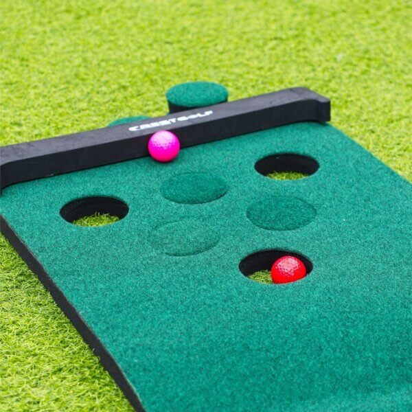 3 our of 6 holes on a golf pong mat are filled in. a Red ball is sitting inside an hole and pink ball sits against the backboard.