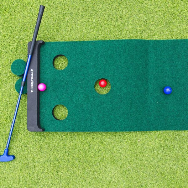 3 our of 6 holes on a golf pong mat are filled in. a Red ball is sitting inside an hole and pink ball sits against the backboard. A blue ball sits away from the holes and a blue adjustable putter is visible on the left