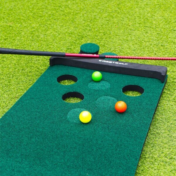 3 our of 6 holes on a golf pong mat are filled in. A green, yellow and orange ball are all sitting on the golf pong mat and not in the holes. A red adjustable putter is visible at the end